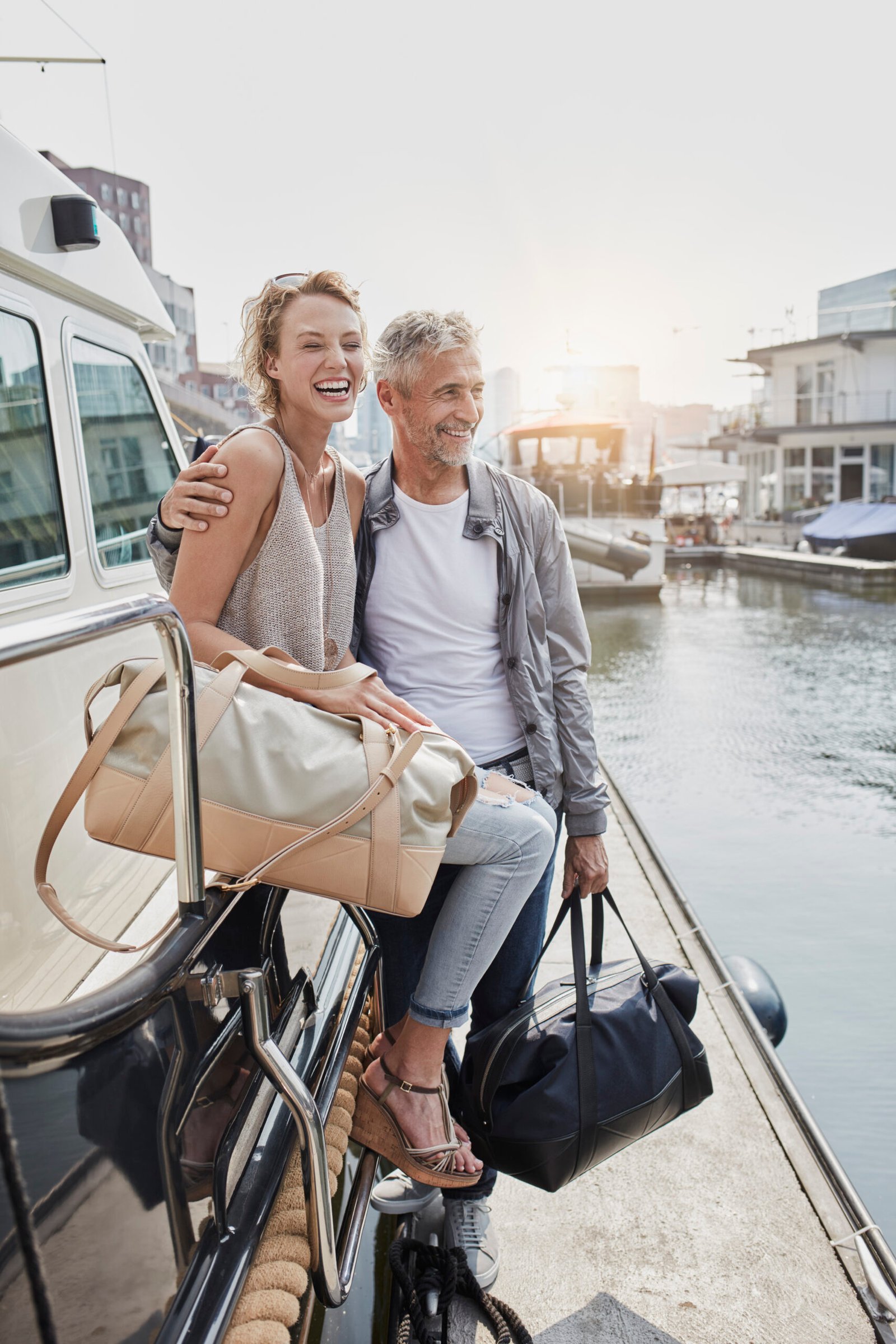 Older man and young woman standing with travelling bags on jetty next to yacht